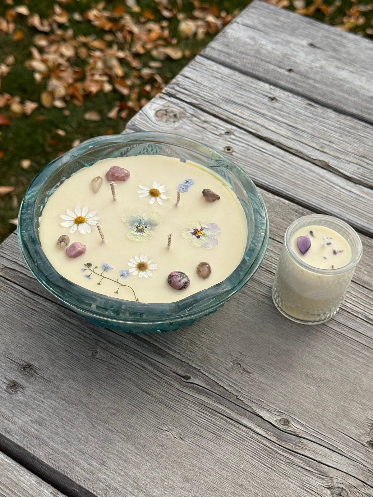 A decorative candle in a round teal glass dish topped with pressed flowers and stones, shown beside a smaller votive candle jar for size.